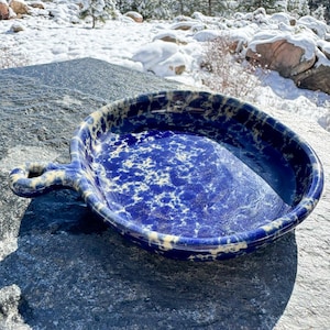 May include: A blue and white speckled ceramic baking dish with a handle. The dish is round with a slightly raised edge and a handle on one side. The background is a snowy outdoor scene.