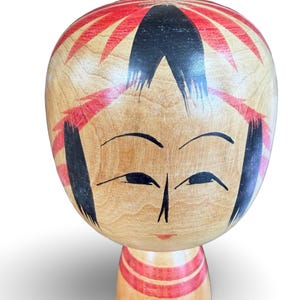 Peut inclure: Une poup&eacute;e kokeshi en bois dot&eacute;e d'une grande t&ecirc;te arrondie avec une finition en bois clair naturel. Le visage est peint &agrave; la main avec des sourcils noirs arqu&eacute;s, des yeux &eacute;troits et une petite bouche rouge. Des d&eacute;tails de cheveux rouges et noirs.