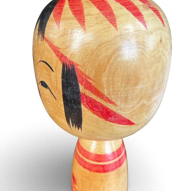 Peut inclure: Une poup&eacute;e Kokeshi en bois dot&eacute;e d'une grande t&ecirc;te arrondie avec des cheveux noirs peints &agrave; la main et des accents floraux rouges. Le visage pr&eacute;sente des lignes noires simples pour les yeux et les sourcils. Le corps cylindrique porte des bandes rouges.