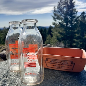 May include: Two clear glass milk bottles with red lettering that reads "Modern Dairy Co. ELGIN ILL" and a rectangular orange bread baker with text that reads "THE ORIGINAL SUFFOLK BREAD BAKER". The items are on a stone surface with a forest background.
