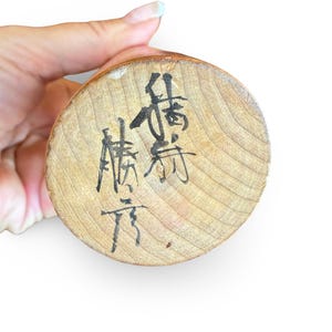 Peut inclure: Une base circulaire en bois clair avec un motif de grain visible, comportant des caract&egrave;res calligraphi&eacute;s &agrave; l'encre noire &eacute;crits verticalement et horizontalement sur la surface. Le bois a un bord arrondi lisse et une couleur beige naturelle.