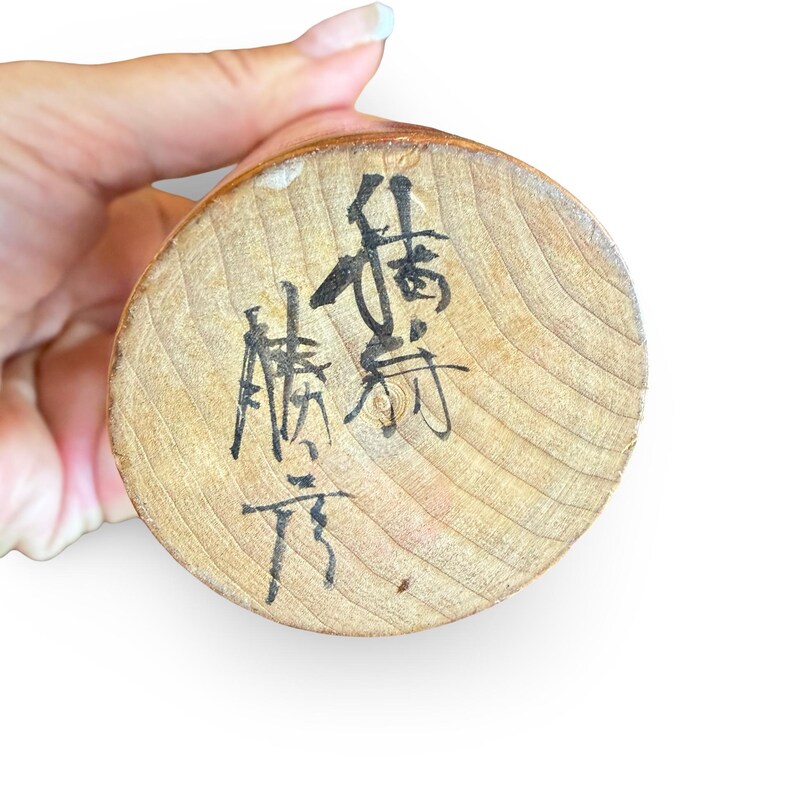 Peut inclure: Une base circulaire en bois clair avec un motif de grain visible, comportant des caract&egrave;res calligraphi&eacute;s &agrave; l'encre noire &eacute;crits verticalement et horizontalement sur la surface. Le bois a un bord arrondi lisse et une couleur beige naturelle.