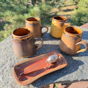May include: Three ceramic mugs with brown and orange glazed rims and a rectangular ceramic tray featuring a metallic tea infuser. The mugs have cylindrical bodies with handles, while the tray has a smooth, glossy finish in shades of orange.