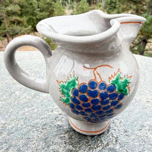 May include: A light gray ceramic pitcher featuring a hand-painted cluster of blue grapes with green leaves and orange outlines. The pitcher has a rounded body, a curved handle, and a wide, shaped rim with a pouring spout on the side.