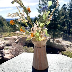 May include: A beige ceramic vase with vertical lines, holding a bouquet of colorful artificial flowers. The flowers include orange, blue, and white blooms, with green leaves and stems. The vase is on a stone surface, with a scenic outdoor background.