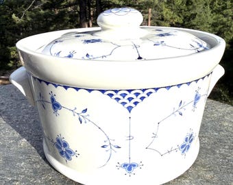 Pode incluir: Uma caçarola de cerâmica branca com tampa, apresentando padrões florais azuis, motivos delicados de videiras e um design de borda azul festonada. O recipiente redondo inclui duas alças laterais e uma tampa a condizer com um puxador.
