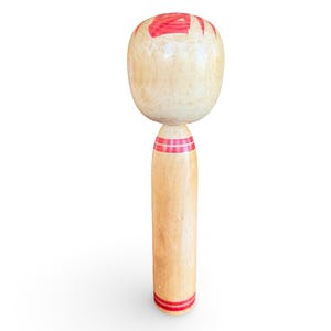 Peut inclure: Une poup&eacute;e Kokeshi en bois clair dot&eacute;e d'une grande t&ecirc;te arrondie avec des motifs floraux peints en rouge sur le dessus. Le corps cylindrique pr&eacute;sente une finition en bois naturel lisse avec deux jeux de rayures rouges horizontales au cou et &agrave; la base.