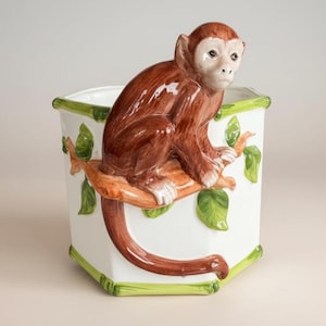 May include: A white hexagonal planter with a brown monkey perched on a branch. The monkey has a light face and brown fur. The planter is trimmed with green bamboo-like accents and green leaves.