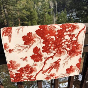 May include: A rectangular cream-colored table runner with a red toile pattern featuring grapes, leaves, birds, and a bee. The runner is draped over a wooden railing, with a blurred natural background.