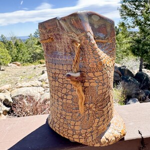 May include: A cylindrical ceramic vessel with a textured, tan-colored surface featuring an embossed floral and grid pattern. The top rim is glazed in a dark brown tone, and a thick, light brown rope is threaded through the upper section as a closure.