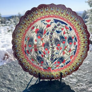 May include: A decorative ceramic plate with a scalloped edge. The plate features a floral design with red, blue, and green accents on a white background. The rim is a dark maroon color. The plate is displayed on a black metal stand.