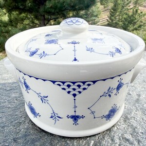 May include: A white ceramic casserole dish with a lid, featuring a blue hand-painted floral pattern and scalloped decorative accents. The round vessel includes two side handles and a matching lid with a central knob, all in a glossy finish.
