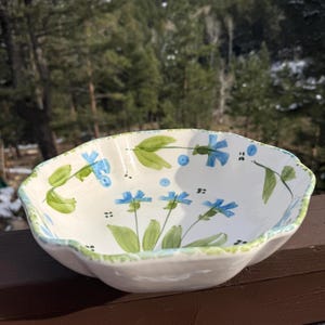 May include: White ceramic serving bowl with a scalloped edge, hand-painted with blue flowers and green leaves. The bowl has a light blue and green trim. A decorative piece for serving salads or displaying fruit.