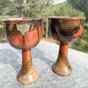 May include: Two handcrafted ceramic goblets featuring a rounded bowl and a flared base. The surface displays a reactive glaze with a blend of deep terracotta red, dark brown, and muted sage green tones, characterized by a distinct drip pattern.
