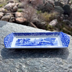May include: A rectangular, blue and white ceramic serving tray with a landscape scene. The tray has a detailed floral border and a central image of a pastoral scene with trees, buildings, and figures. The tray is approximately 12 inches long.