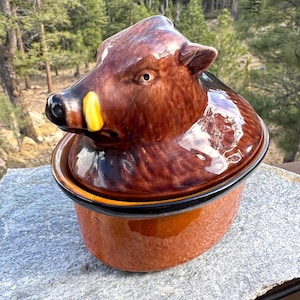 May include: A ceramic boar-shaped casserole dish with a lid. The dish is primarily brown with a black rim and a yellow accent on the boar's snout. The lid is shaped like the boar's head.