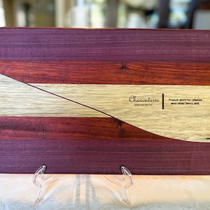 May include: A rectangular wooden charcuterie board with a unique design. The board features alternating stripes of purple, red, and light wood, with a diagonal cut across the top. Text on the board reads "Charcuterie" and "French word for cheese and other fancy sh*t."