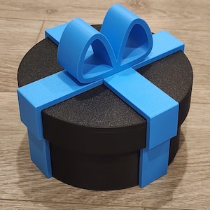 Op de afbeelding: Een ronde zwarte container met een matte afwerking, voorzien van een felblauw decoratief lint en een strikelement op het deksel. Het blauwe lint loopt langs de zijkanten van de cilindrische doos, wat zorgt voor een cadeau-achtig uiterlijk.