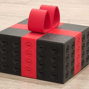 May include: A square box constructed from black and red interlocking building blocks, featuring a red ribbon and bow on top. The sides of the box are textured with a grid of circular studs, and the central red band also displays a vertical row of studs.