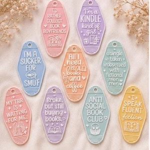 Op de afbeelding: Een verzameling van negen ruitvormige plastic sleutelhangers in pastelkleuren, elk voorzien van witte reliëftekst en illustraties gerelateerd aan lezen. De sleutelhangers bevatten teksten zoals I'd rather collect book boyfriends, I'm a Kindle kind of girl, I'm a sucker for smut, All I need is books and coffee, single taken obsessed with fictional men, My TBR is waiting for me, Broke but still buying books, Anti social book club en I speak fluent fiction.