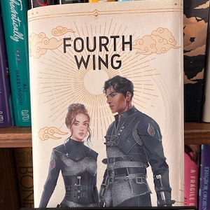 May include: A hardcover book titled Fourth Wing with a cream-colored cover featuring an illustration of two figures in black tactical gear. Below the book, a black rectangular shelf sign with gold-colored text reads Shelf Royalty.