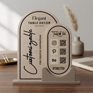 以下が含まれることがあります： アーチ型のトップを持つ明るい色の木製看板。Elegant TABLE DECORおよびCustomizable FOR CREATIVE INSPIRATIONという文字が記されています。前面に取り付けられた小さな楕円形の看板には、TablEtchのロゴ、ソーシャルメディアのアイコン、QRコードが表示されています。