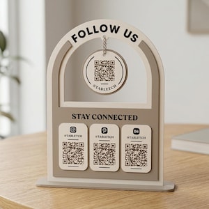 Può includere: Un espositore ad arco color beige e marrone chiaro con il testo FOLLOW US e STAY CONNECTED. Un'etichetta circolare con un codice QR e il testo @TABLETCH pende dall'arco. Tre etichette rettangolari sottostanti mostrano icone e codici QR.