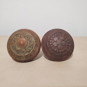 May include: Two circular metal door knobs with a weathered patina. The left knob features a raised central dome surrounded by intricate swirling scrollwork. The right knob displays a dark, textured surface with a central star-like emblem.