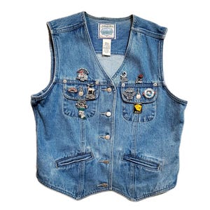 May include: Light blue denim vest with multiple decorative pins. The vest has a V-neck, button closure, and two front pockets. The pins include various designs such as a motorcycle, a deer, and a butterfly. The vest has a label that says "St. John's Bay".