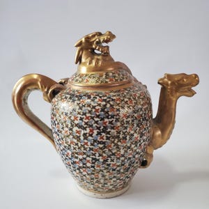May include: An ornate ceramic teapot with a dragon-shaped spout and lid finial. The teapot features a gold handle and spout, and a patterned body with blue, red, and black cross-shaped designs. The teapot is a decorative item.