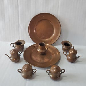 May include: A collection of antique copper miniature dishware, including plates, pitchers, and teapots. The set features a warm, reddish-brown color and is arranged on a white surface. The word "CHILE" is stamped on the plates.
