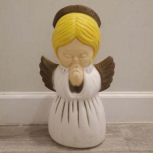 May include: A vintage Christmas angel figurine with a halo, praying hands, and wings. The angel has yellow hair, a white robe, and brown wings. The base of the figurine has star-shaped cutouts. This decorative angel is a classic holiday decoration.
