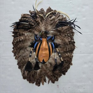 May include: A decorative mask featuring a tan face with black and blue painted markings. The mask is surrounded by a dense layer of brown, black, and cream-colored feathers. Two beaded strands with black feathers hang from the sides of the mask.