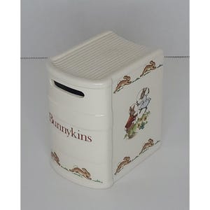 May include: A white ceramic Bunnykins bank shaped like a book. The bank features illustrations of rabbits and the word "Bunnykins" in brown lettering. The top has a ribbed design, and a slot is visible.