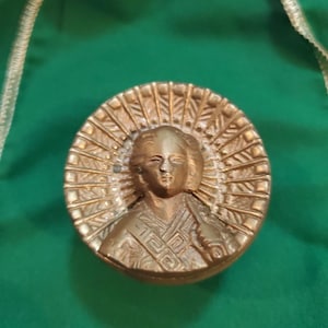 May include: A bronze-colored, circular medallion featuring a detailed relief of a person with a halo. The figure is depicted with stylized hair and clothing. The medallion is set against a green background.