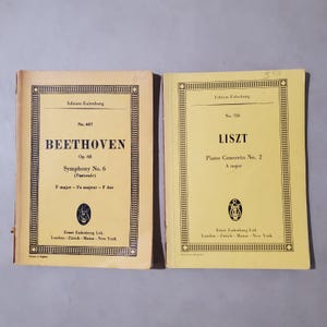 May include: Two vintage Edition Eulenburg music scores with pale yellow covers. The left score is Beethoven Symphony No. 6, Op. 68. The right score is Liszt Piano Concerto No. 2, A major. Both feature black text, decorative borders, and publisher logos.