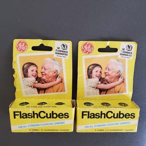 May include: Two vintage yellow FlashCubes boxes. Each box features a photo of a young girl hugging an older man. The boxes have the GE logo and text that reads "FlashCubes" and "For All Standard Flashcube Cameras". Each box contains 3 cubes and 12 guaranteed flashes.