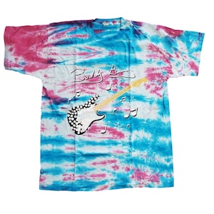 Peut inclure: Un t-shirt tie-dye aux couleurs rose, bleu et blanc. Le t-shirt présente le texte "Buddy Ace" au-dessus d'un dessin de guitare avec des notes de musique. La guitare a un motif à pois noirs et blancs.