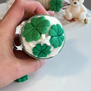 May include: A glass jar candle topped with white wax whipped to resemble frosting, decorated with three green four-leaf clover wax embellishments. The candle features a central wick and is viewed from directly above, showcasing the textured wax.