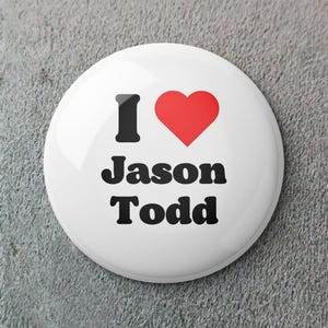 Puede incluir: Un botón blanco con el texto "I ❤️ Jason Todd" en negro y un símbolo de corazón rojo. El botón es redondo y tiene un acabado brillante, ideal para adjuntar a la ropa o las bolsas.