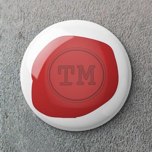 Puede incluir: Un botón blanco con una forma roja brillante e irregular. Dentro de la forma roja hay un diseño circular con las letras "TM" en negrita, sin serifa. El botón está sobre un fondo gris texturizado.