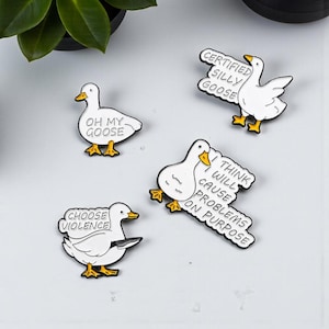 Könnte beinhalten: Vier weiße Gänse-Emaille-Anstecknadeln mit schwarzen Umrissen und gelben Füßen und Schnäbeln. Jede Anstecknadel hat einen anderen Spruch: "OH MY GOOSE", "CHOOSE VIOLENCE", "CERTIFIED SILLY GOOSE" und "I THINK WILL CAUSE PROBLEMS ON PURPOSE".