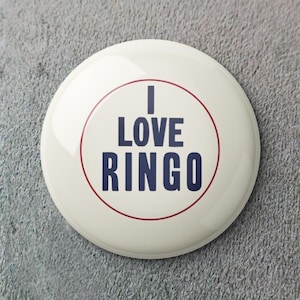 Puede incluir: Un botón redondo blanco roto con el texto "I LOVE RINGO" en azul marino, rodeado por un fino círculo rojo. El botón tiene un acabado brillante y está sobre un fondo gris texturizado.