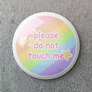 Puede incluir: Un botón redondo de colores pastel con un diseño de arcoíris y el texto "please do not touch me" en rosa. El botón presenta acentos de estrellas y un borde blanco. La estética general es linda y caprichosa.