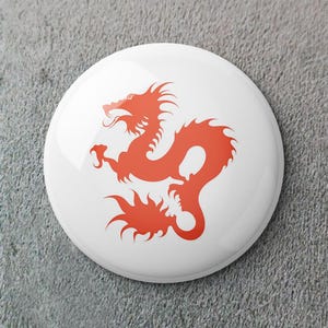 May include: A white button with a red dragon design. The dragon is stylized with a curved body, sharp claws, and spiky details. The button has a glossy finish and is set against a gray background.