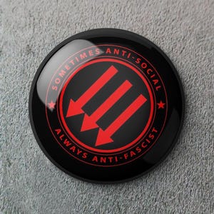 May include: A black circular button with red text and arrows. The text reads "SOMETIMES ANTI-SOCIAL" and "ALWAYS ANTI-FASCIST" around the edge. Three red arrows point downward in the center.