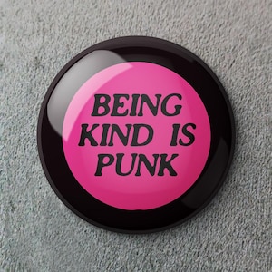 May include: A round button with a glossy finish. The button has a black border and a bright pink center. Bold black text in the center reads "BEING KIND IS PUNK". The button is set against a textured gray background.