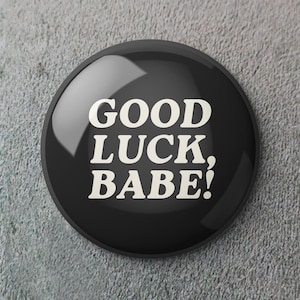Może przedstawiać: Błyszczący, czarny guzik z białym napisem "GOOD LUCK, BABE!". Okrągły guzik ma odblaskową powierzchnię i jest umieszczony na szarym, teksturowanym tle.
