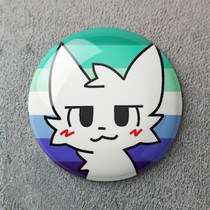 Puede incluir: Un botón redondo con un diseño de cara de gato blanco. El botón presenta un fondo con los colores de la bandera del orgullo gay. El gato tiene ojos negros, una pequeña nariz negra y marcas rojas de rubor en las mejillas.