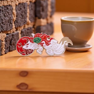 Può includere: Una spilla smaltata dettagliata raffigurante una volpe bianca con dettagli rossi e bianchi e un design circolare rosso. La spilla ha un contorno dorato e un accento circolare verde. La spilla è su una superficie di legno.